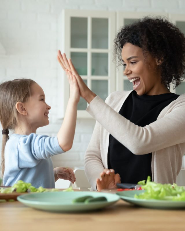 Overjoyed young African American mom and little Caucasian daughter give high five cooking at home together, smiling biracial mother and small girl child have fun preparing healthy food in kitchen
